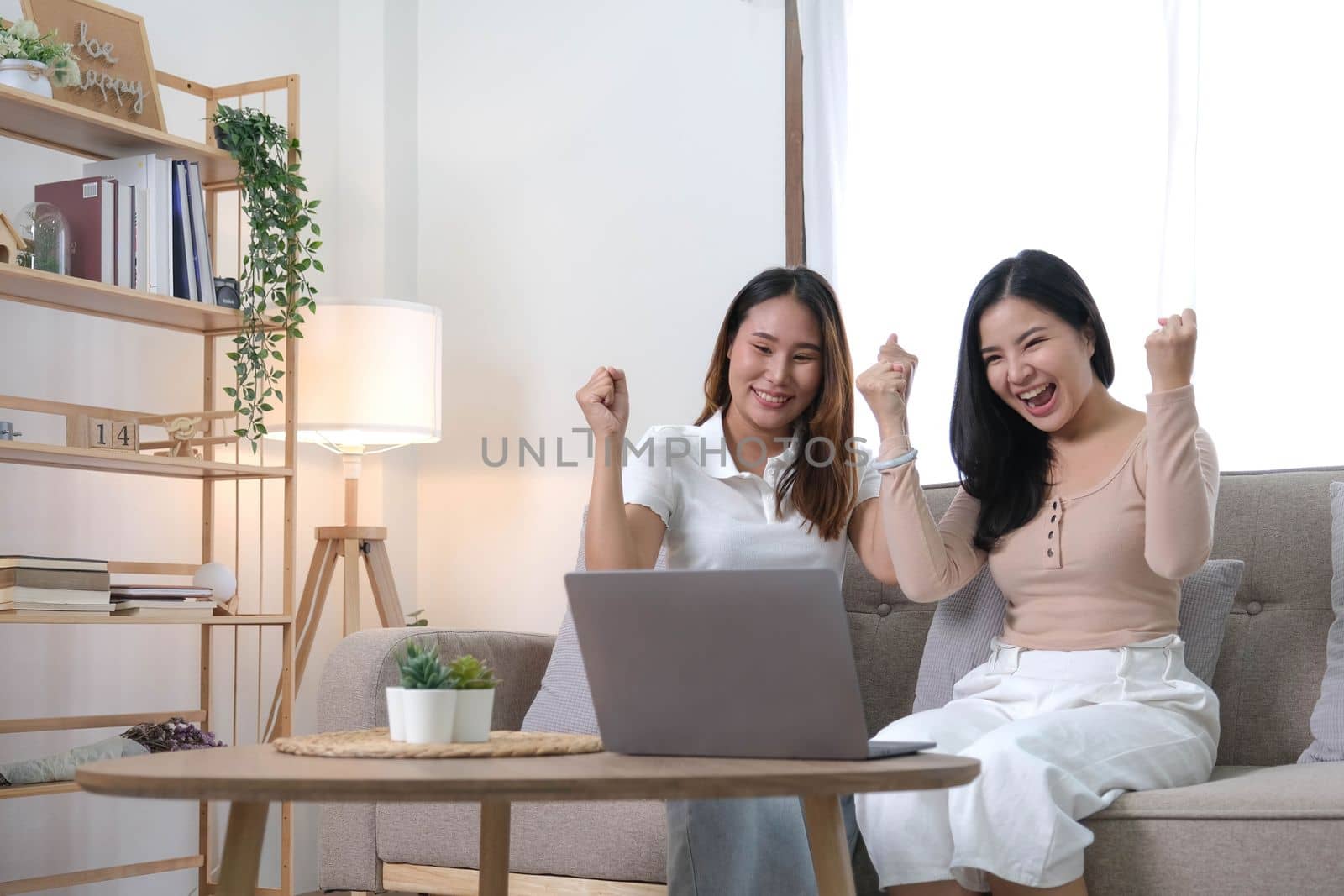 Two happy Asian women best friends in casual wear laughing while working with laptop at home in living room by wichayada