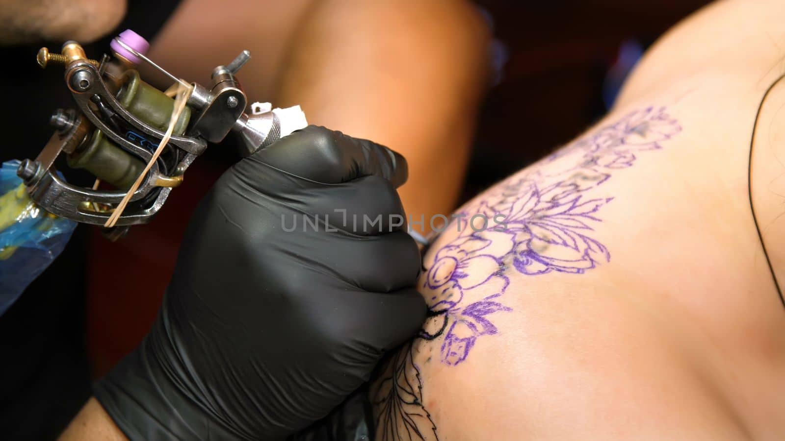 close-up, in a tattoo salon , a specialist is doing a tattoo on woman's back, black paint floral ornament. a man works in special black gloves, on special equipment. by djtreneryay