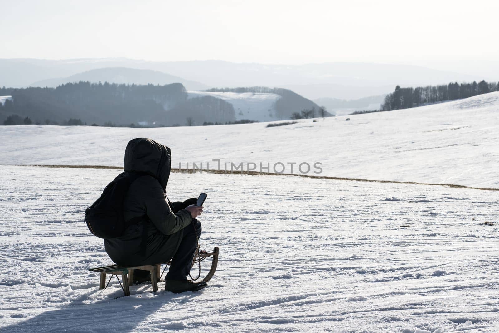 a man looks at his mobile phone sitting in the cold on top of a snowy mountain in winter on a children's sled wrapped in his winter warm jacket by Costin