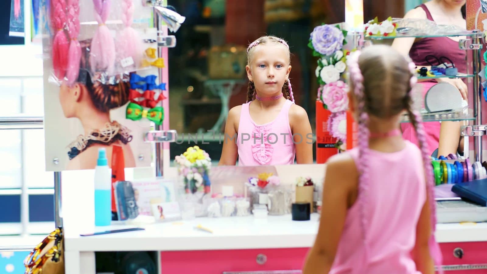 Beautiful blond girl, of seven years old, braided two pigtails, do a hairstyle with pink locks of hair in a beauty salon, a hairdresser's salon, in front of a large mirror. a little princess. High quality photo