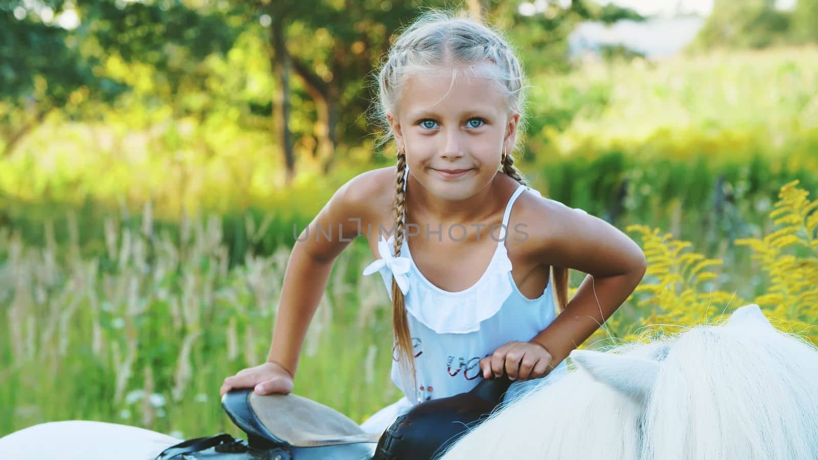 Blond girl of seven years, in a white dress, climbs, sits on a pony. Cheerful, happy family vacation. Outdoors, in the summer, near the forest. High quality photo
