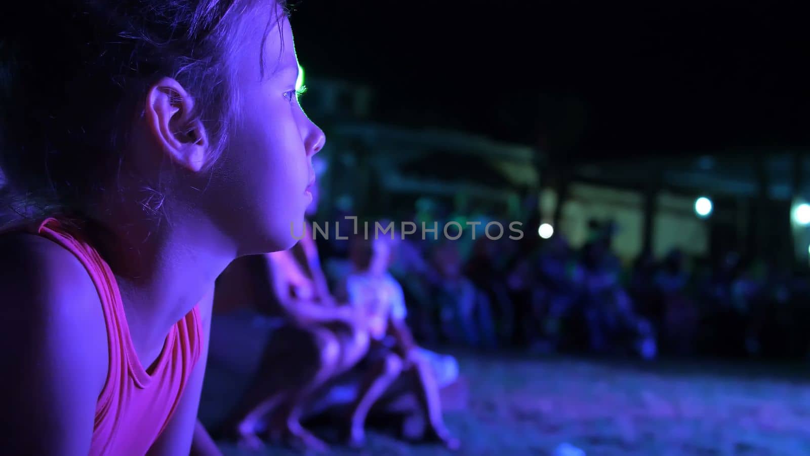 summer, night, in the rays of soffits, a girl of seven years old, watching the night concert in an open-air theater, on vacation. many children are in the background. by djtreneryay