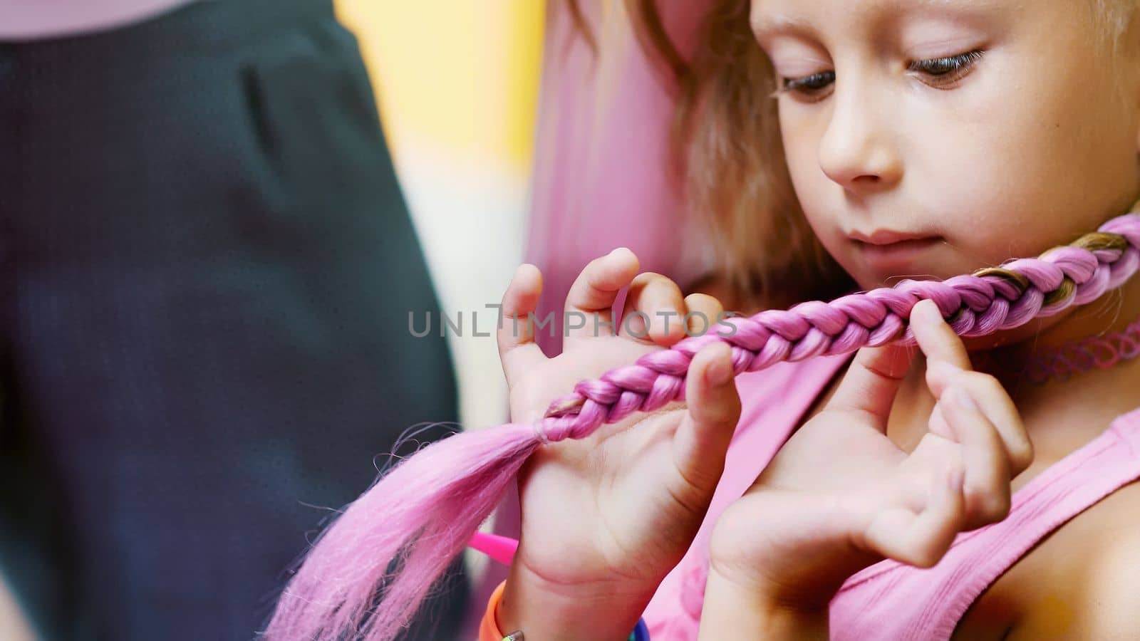 Beautiful blond girl, of seven years old, braided two pigtails, do a hairstyle with pink locks of hair in a beauty salon, a hairdresser's salon, in front of a large mirror. a little princess. High quality photo