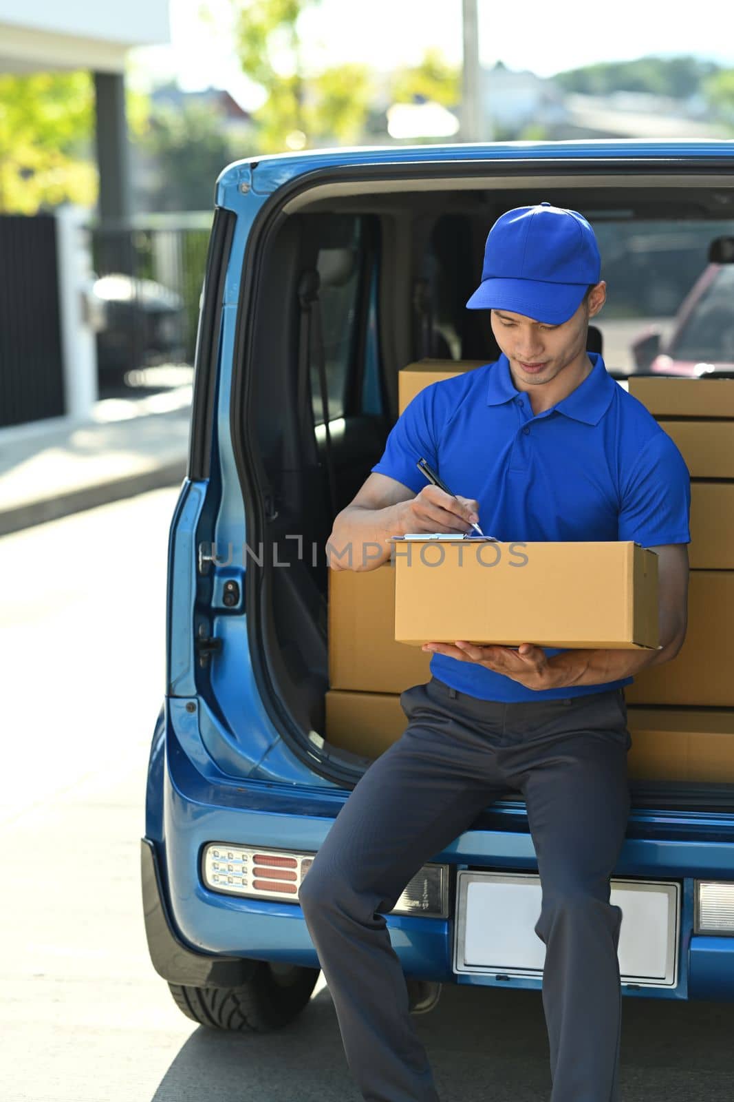 Man courier in blue uniform sitting in open delivery van and checking checking address details of client on clipboard. 