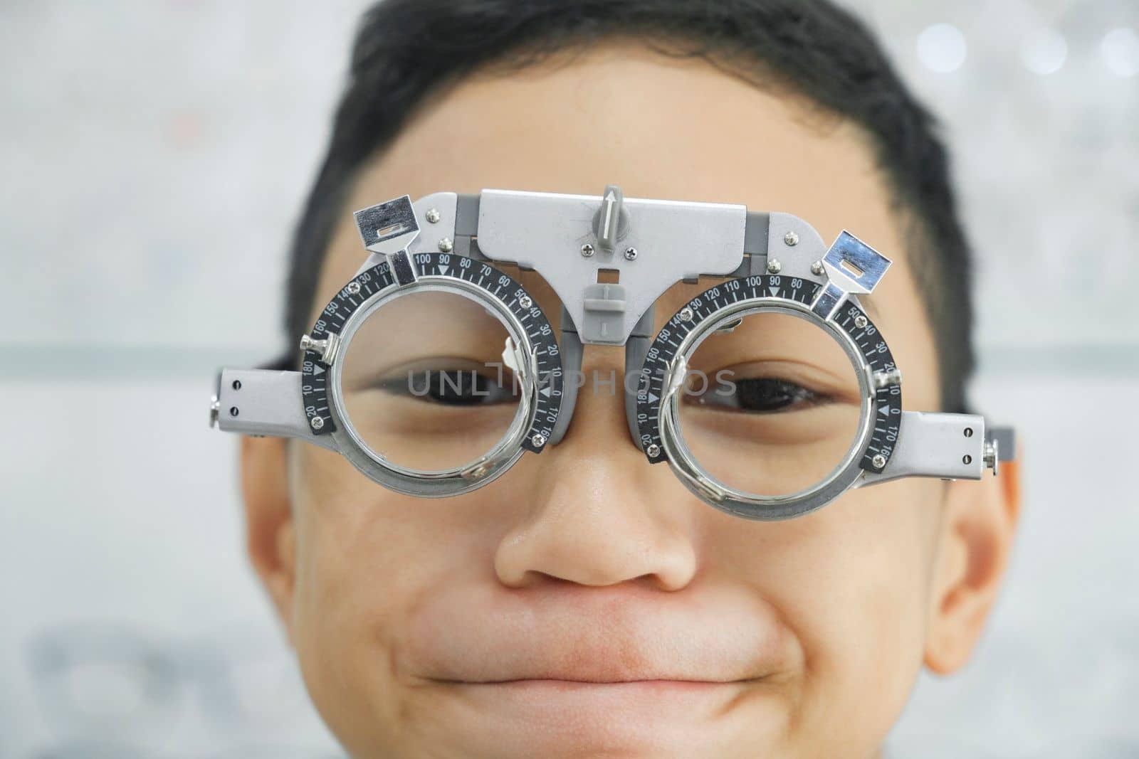 Smart young boy sitting in optometrist cabinet having his eyesight checking, examining, testing with trial frame glasses by professional optician for new pairs of eyeglasses.