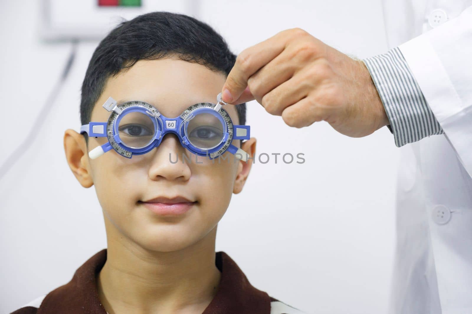 Smart young boy sitting in optometrist cabinet having his eyesight checking, examining, testing with trial frame glasses by professional optician for new pairs of eyeglasses.