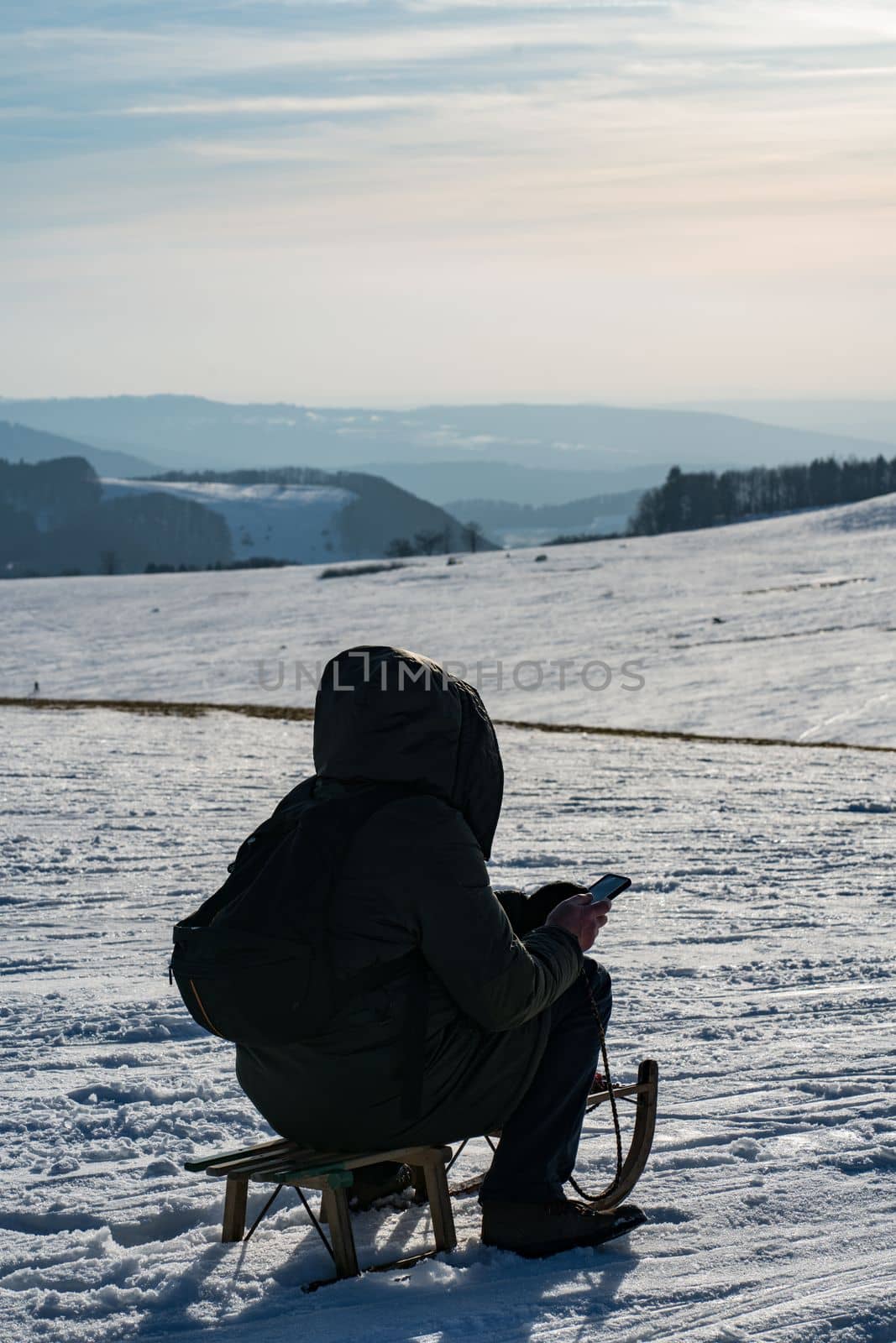 a man looks at his mobile phone sitting in the cold on top of a snowy mountain in winter on a children's sled wrapped in his winter warm jacket by Costin