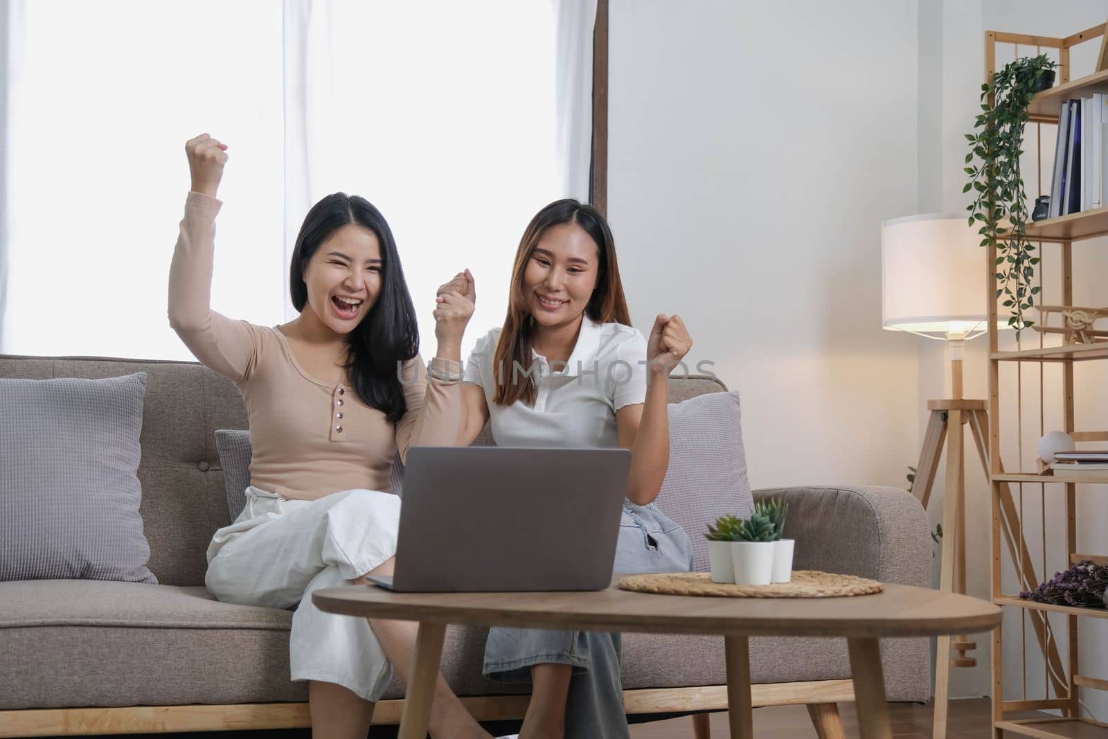 Two happy Asian women best friends in casual wear laughing while working with laptop at home in living room by wichayada