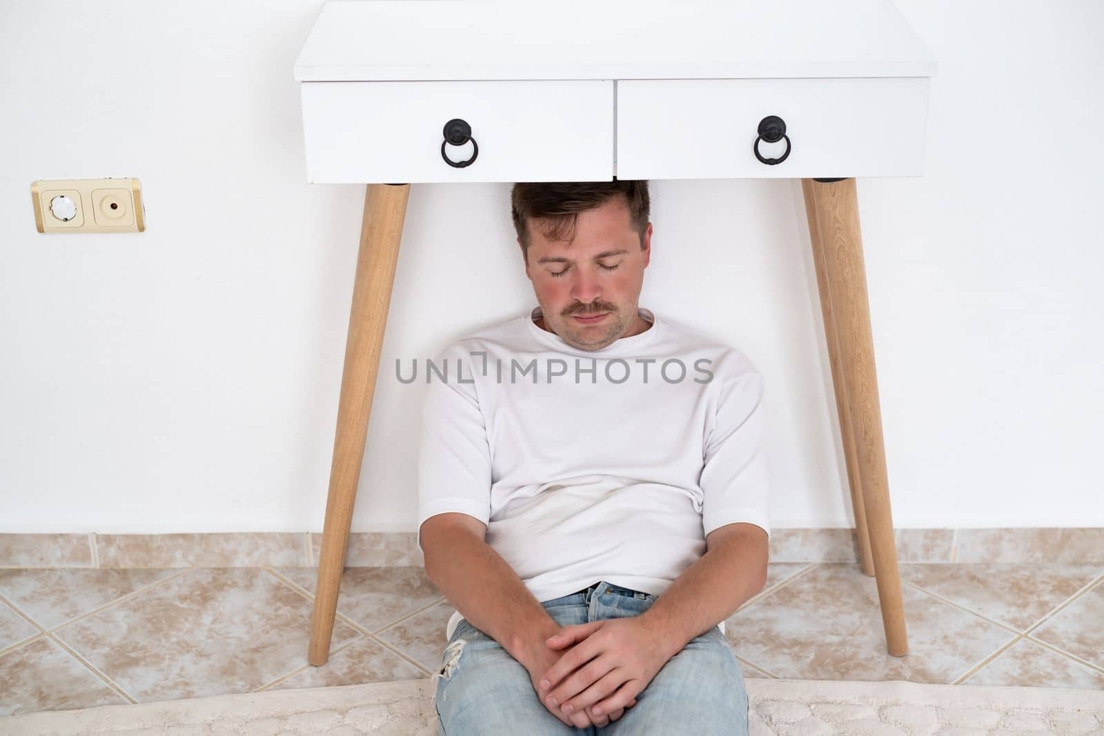 Scared man hiding under table in living room. Live me alone. Stress concept