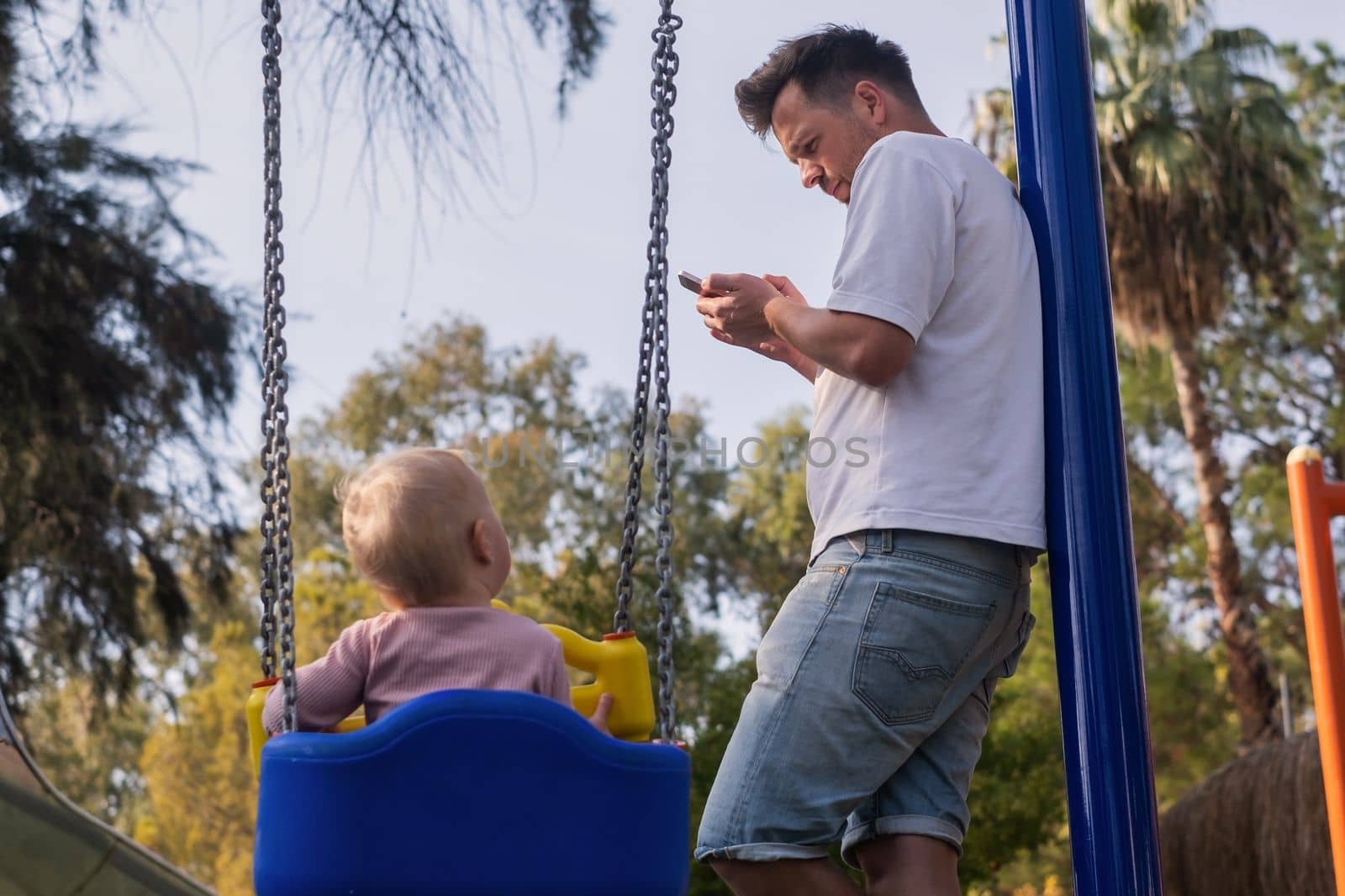 Father walking with child on a playground looking on phone ignoring his baby girl swinging. Phone addiction.