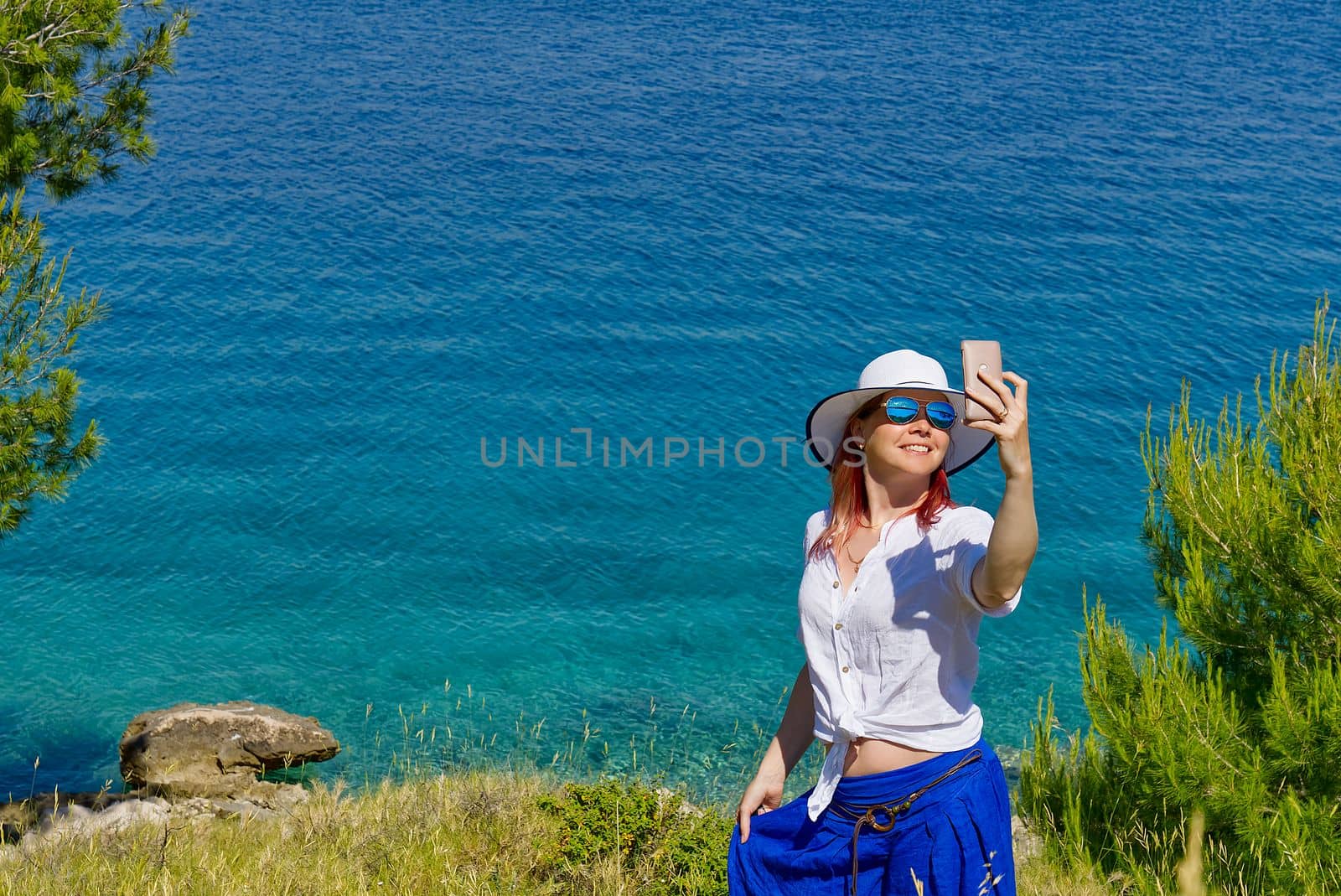 Smiling young woman in sunglasses and straw hat taking selfie against sea and blue sky.