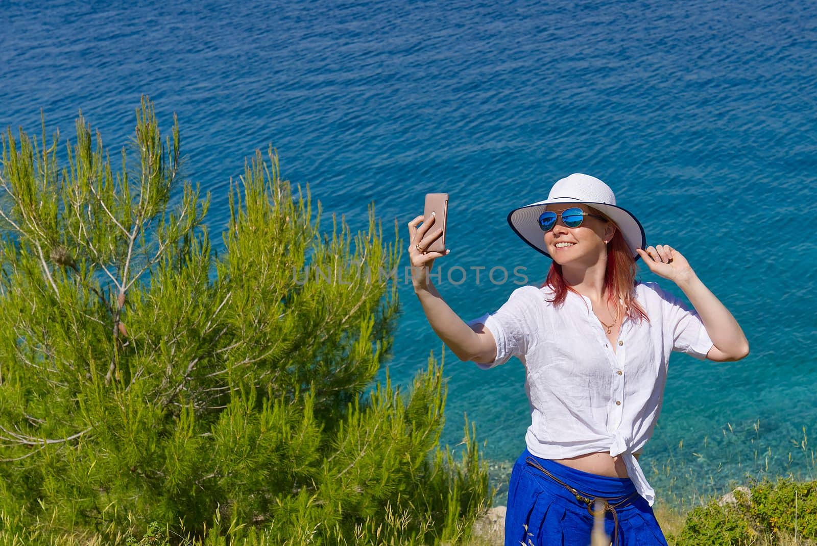 Smiling young woman in sunglasses and straw hat taking selfie against sea and blue sky by PhotoTime