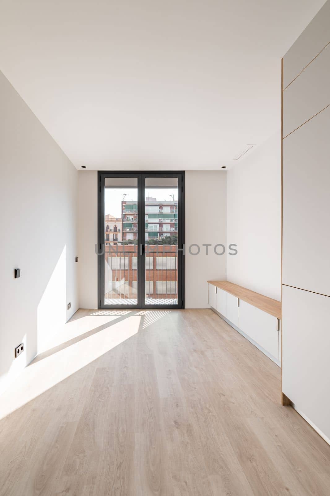 Direct view of black metal framed glass sliding doors at end of an empty bright clean room. Doors leading to spacious balcony overlooking neighboring house. Sunlight enters through glass door