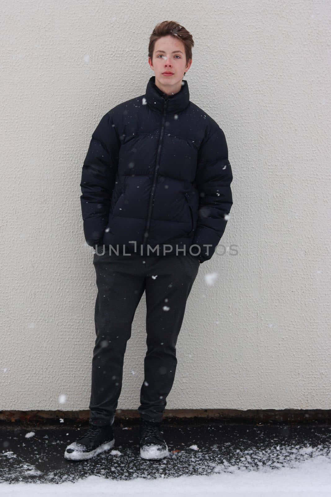 Cute teenage boy at full height in front of a wall in snowy weather. Boy holding hands in pockets during snowfall