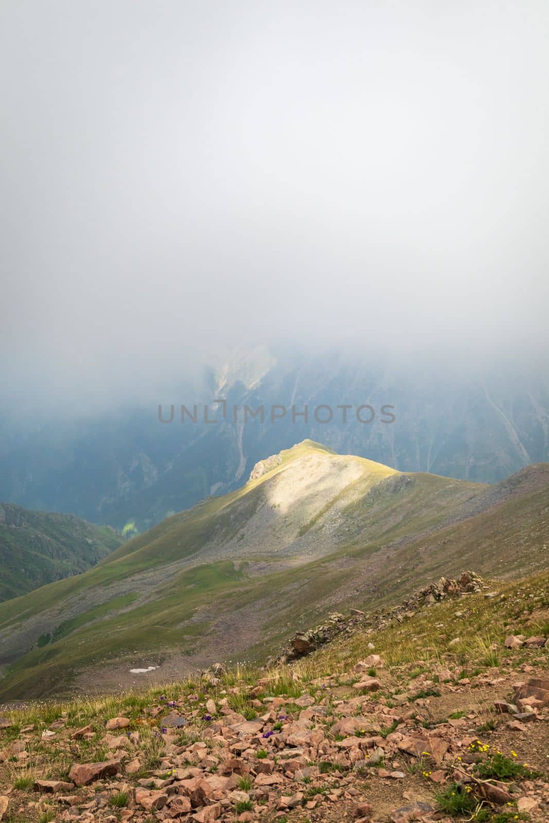 Mysterious minimalist high-altitude picturesque mountain landscape with clouds touching large peaks and a spot of light on a mountain. Copy space, vertical.