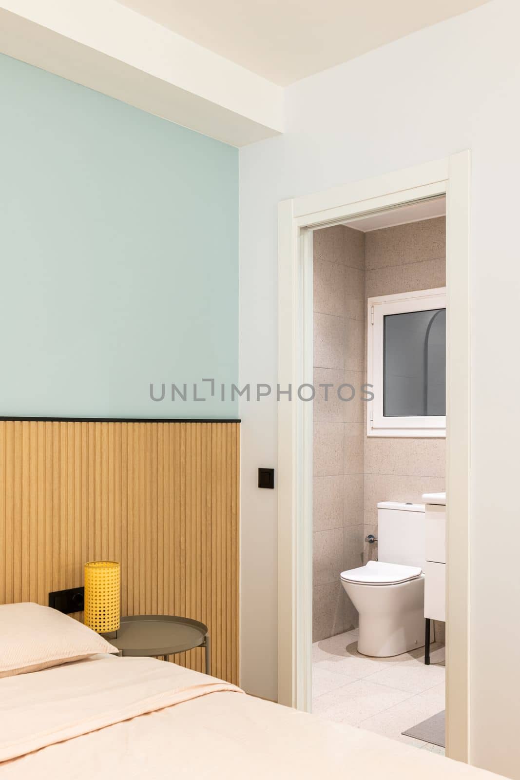 Open door to toilet room from cozy bedroom. Through doorway you can see white toilet bowl, tiled walls and floors, sink edge on black-legged vanity, and frosted plastic window. by apavlin