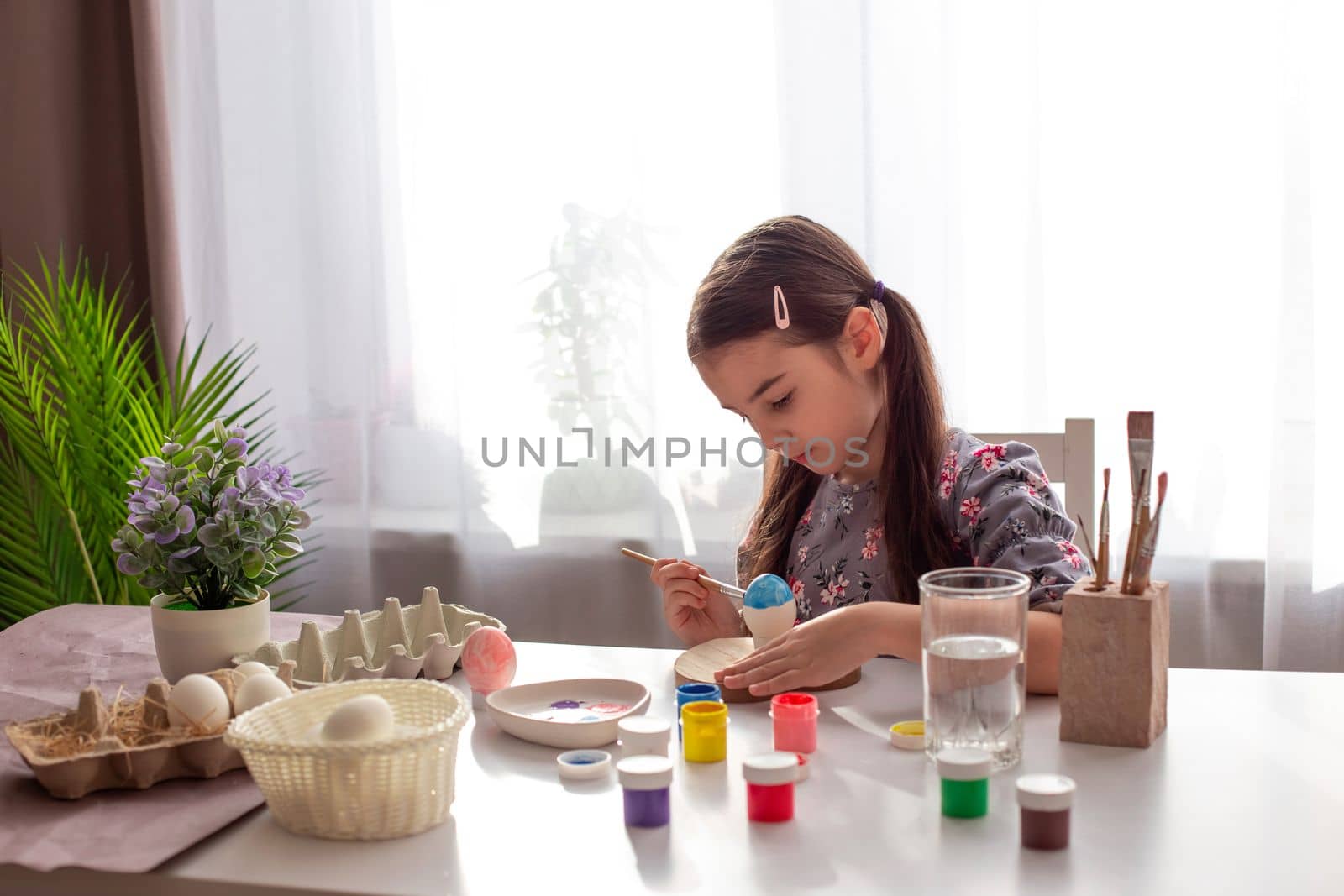 A cute little girl sits at a white table by the window, paints eggs with a brush and paint in jars. Copy space
