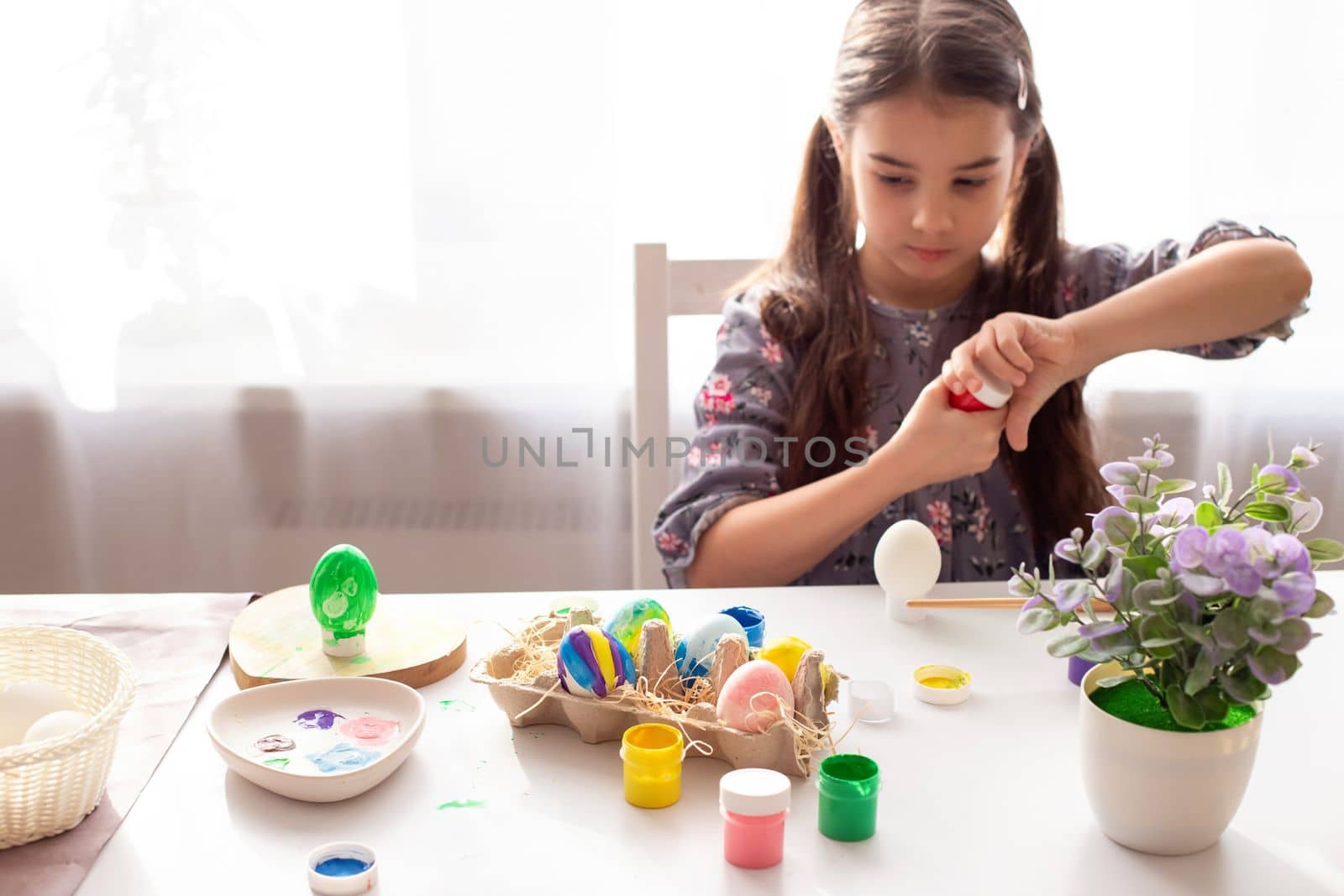 Multi-colored painted eggs in a tray stand on the table, the girl opens a jar of paint in the background. The concept of preparing for the Easter holiday. copy space