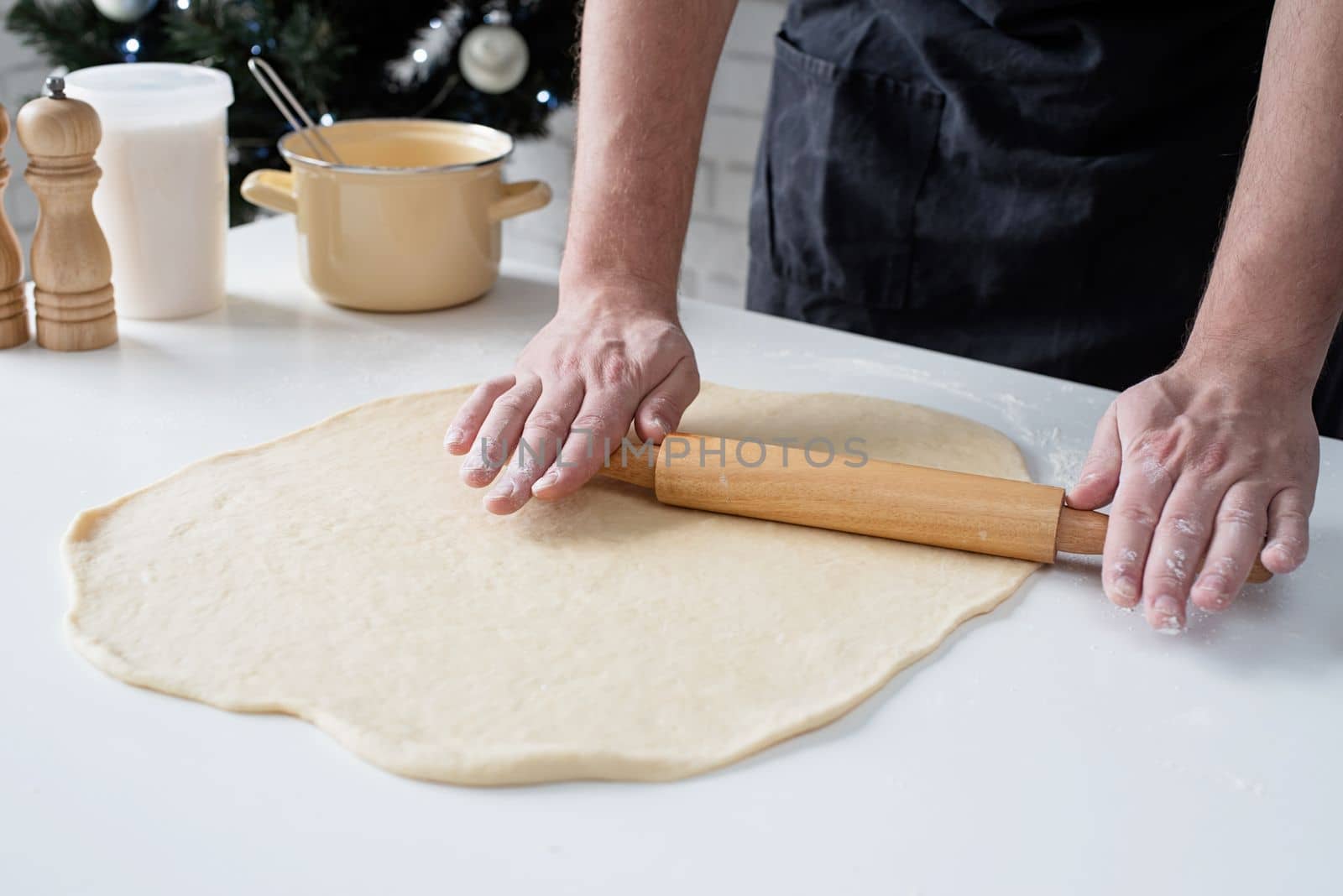 Male chef working with dough, cooking cinnamon rolls, christmas tree with lights on the background, festive cooking