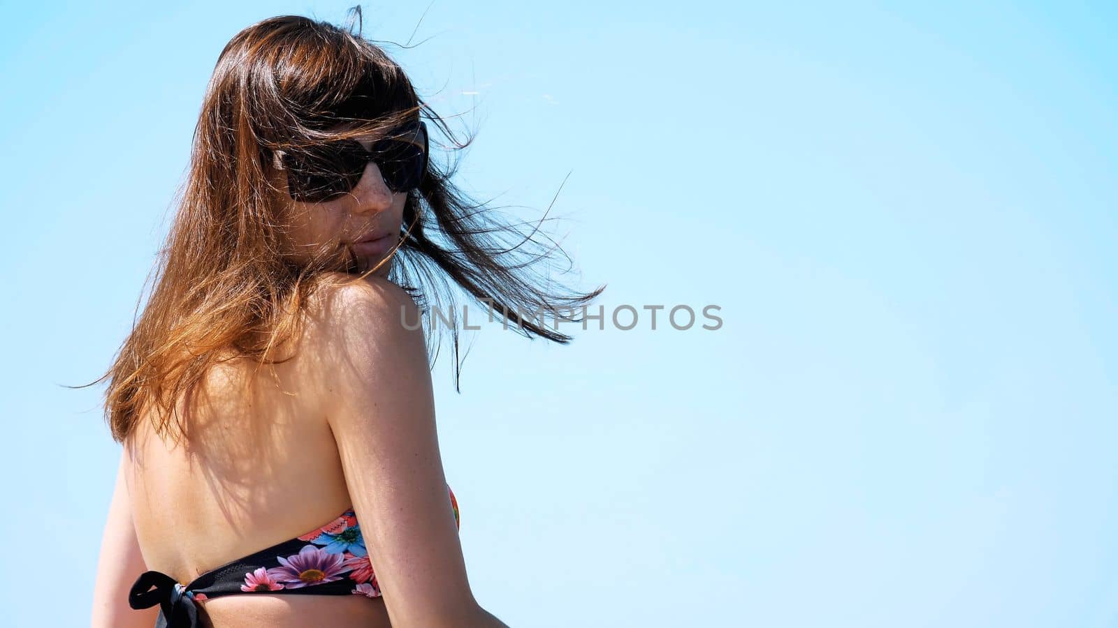 summer, sea, portrait from the back, beautiful young brunette woman wearing a bathing suit and sunglasses, enjoying the rest, her hair are fluttering in the wind. High quality photo