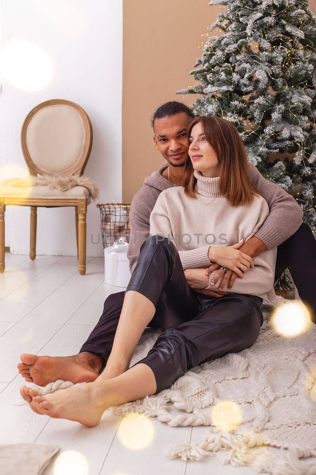 A stylish dark-skinned guy in a brown sweatshirt is sitting hugging his girlfriend, near the Christmas tree in the room. Vertical