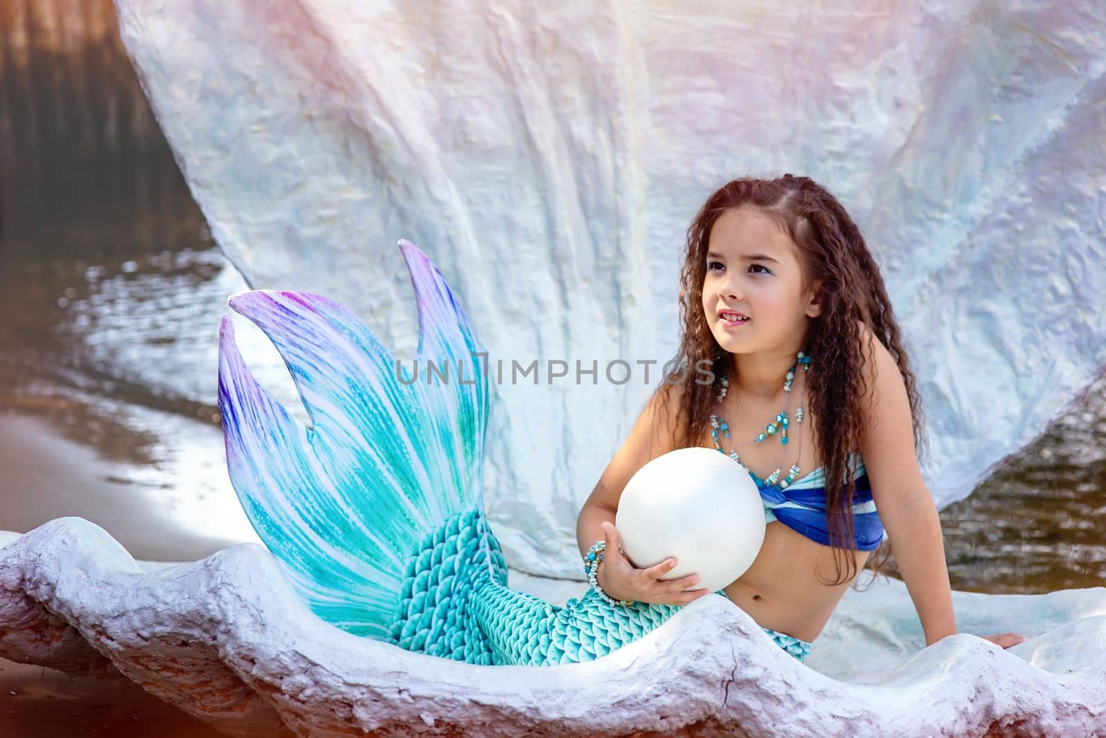 A beautiful happy little brunette girl in a blue mermaid costume, sits in a large sea shell, outdoor, hold a big pearl . Close up. Copy space