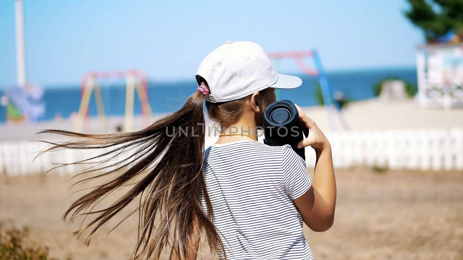 back view, teenage child, long-haired blonde girl in a blazer listening to music with bluetooth portable speaker , on the beach, dancing , on a hot summer day. High quality photo