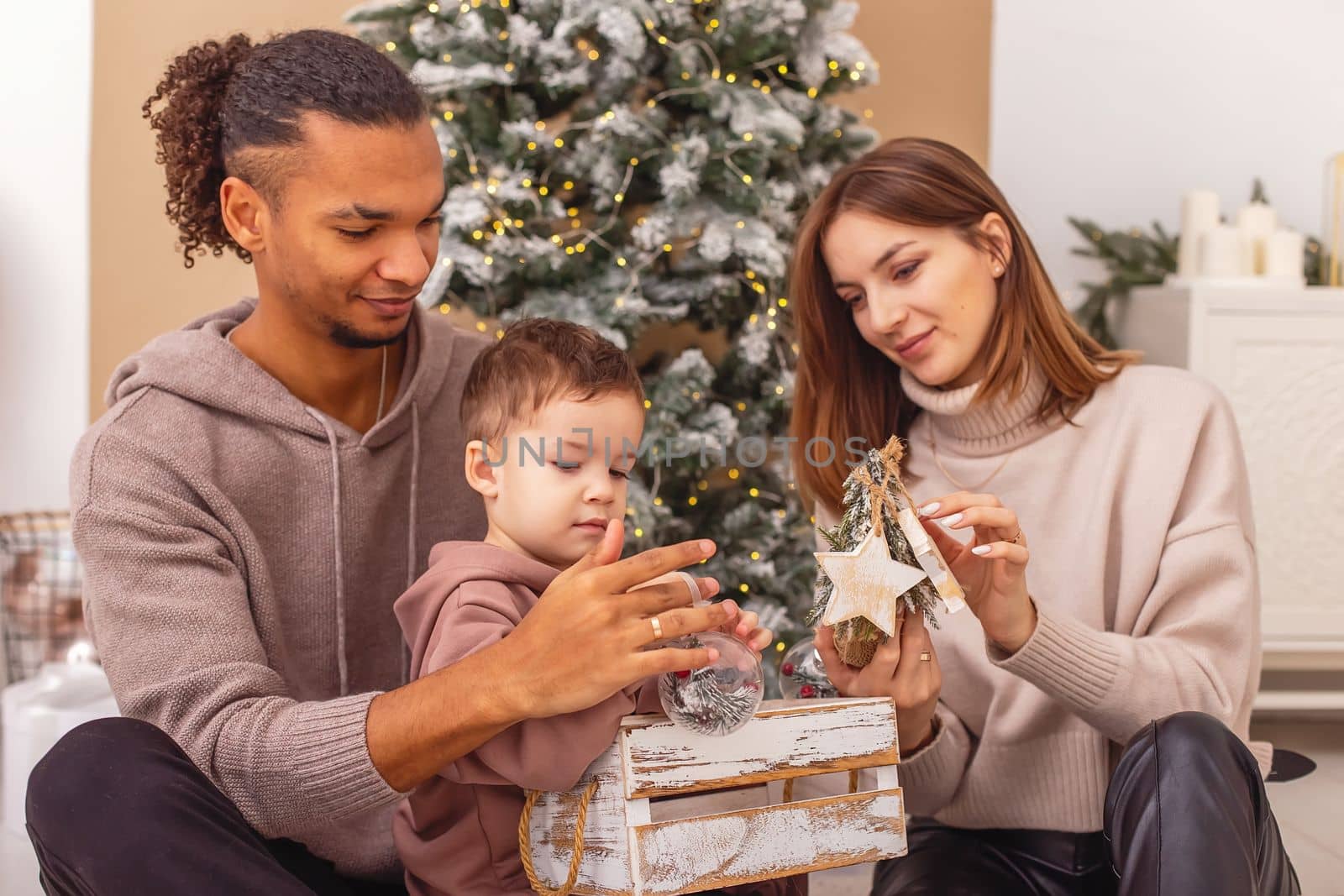 A happy multi-racial family with a little boy is sitting on a knitted blanket, holding a wooden Christmas toys. Close up