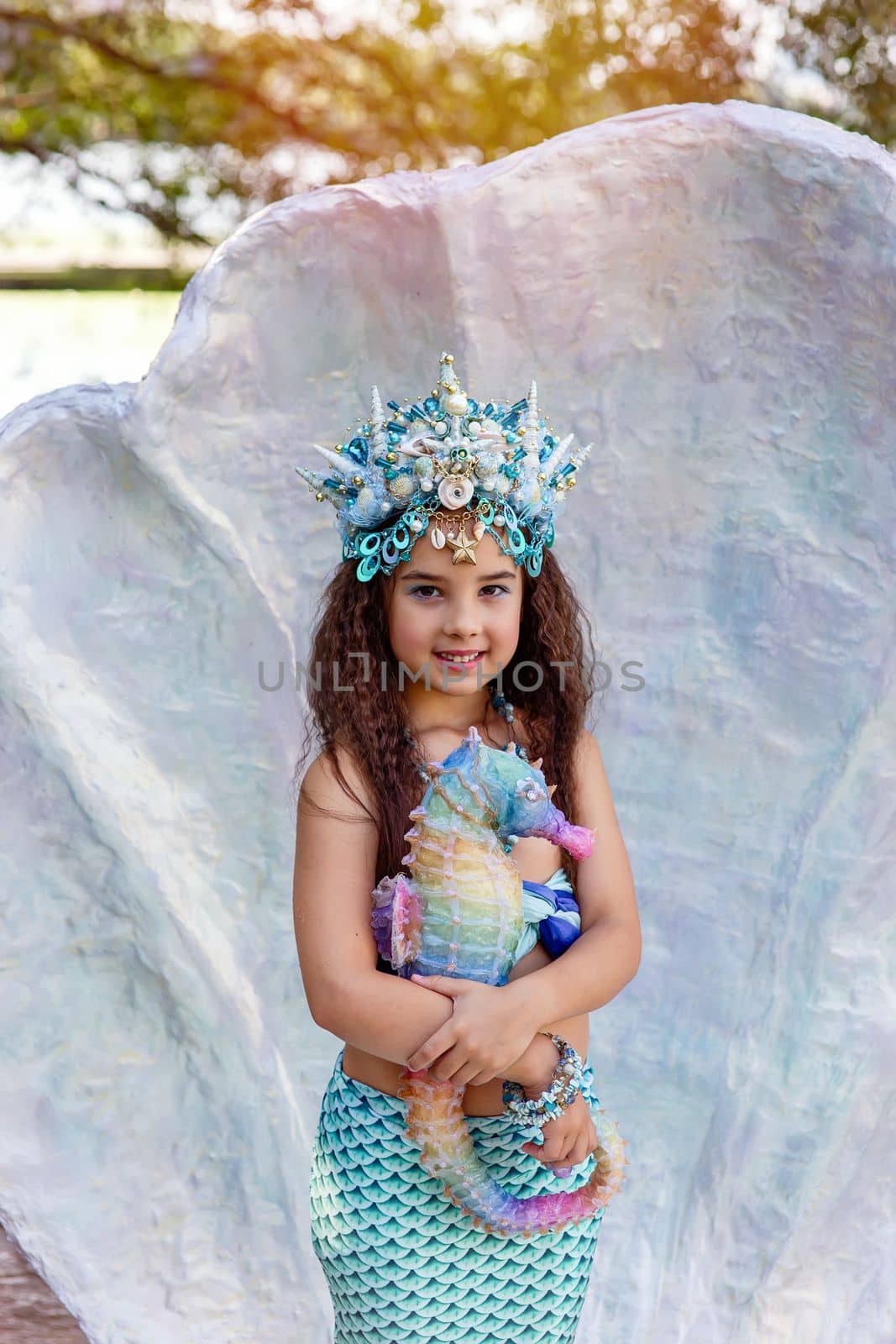 A smiling little girl in a mermaid costume in a large seashell, holding a colorful seahorse toy. by Zakharova