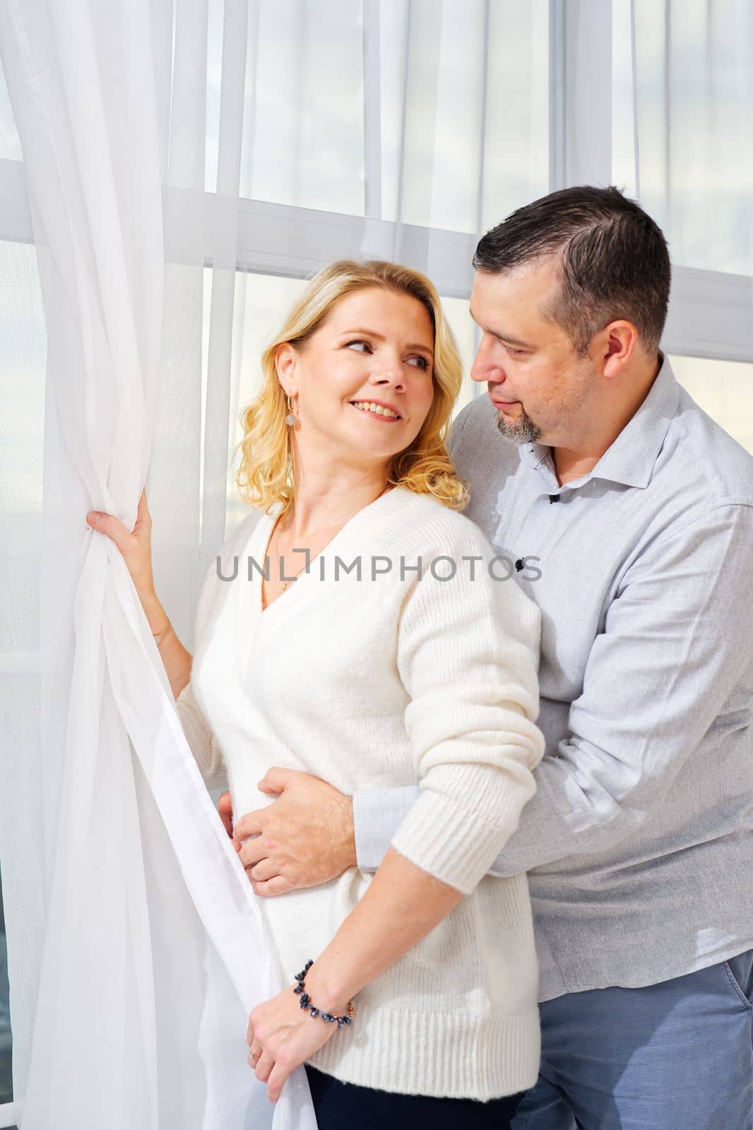 Middle aged blond woman and man portrait. 40s couple embracing in front of window.
