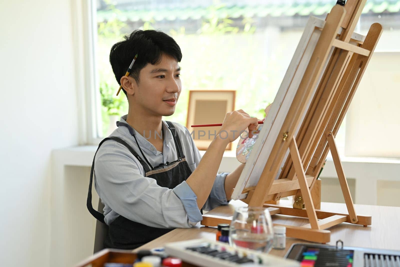 Concentred asian male artist sitting in front of canvas and painting picture with watercolor. Leisure activity concept.