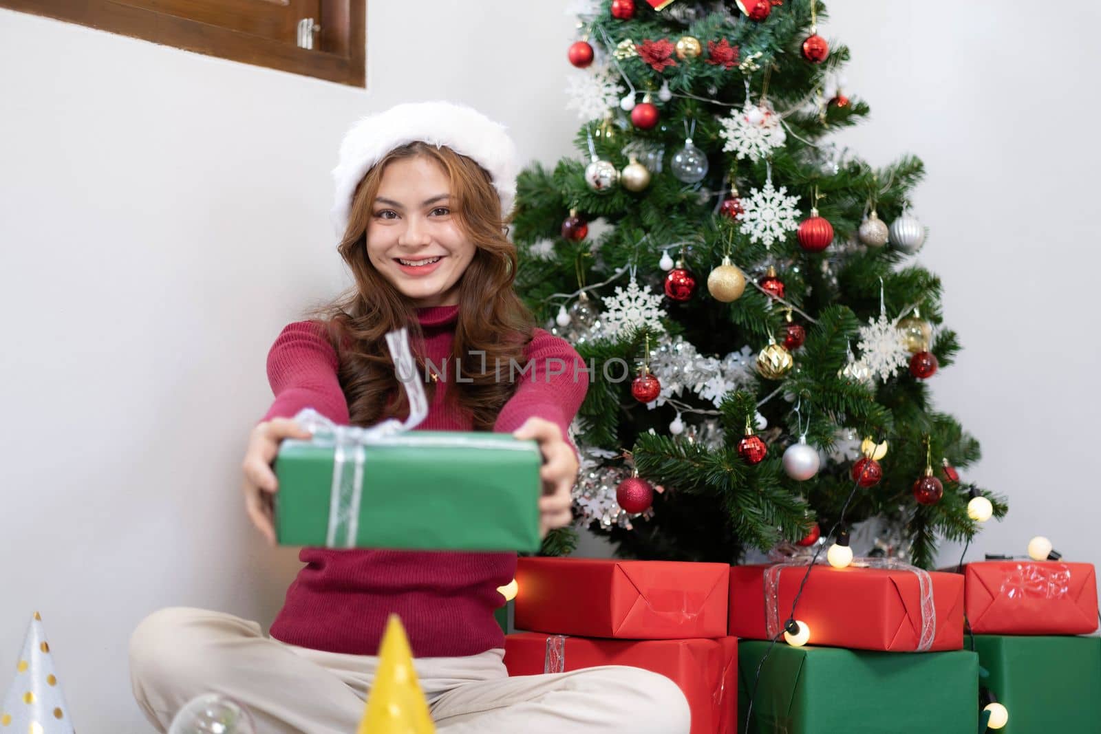 Cheerful asian young beautiful woman wearing santa hat showing open gift box with happy smiling face sitting in living room background...