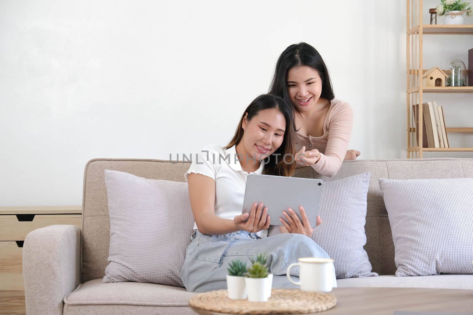 Two happy Asian women best friends in casual wear laughing while working with tablet at home in living room by wichayada