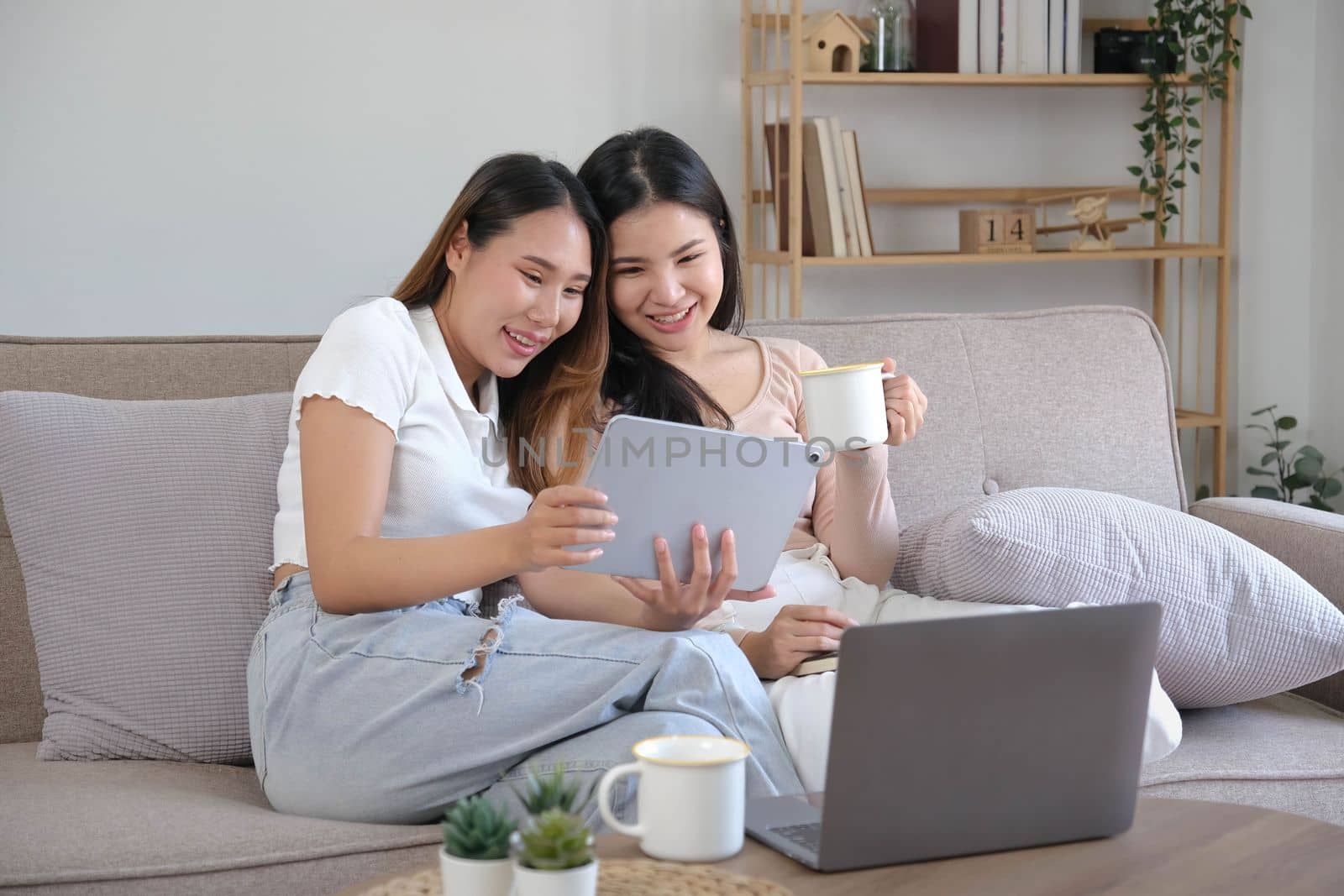 Two happy Asian women best friends in casual wear laughing while working with tablet at home in living room by wichayada