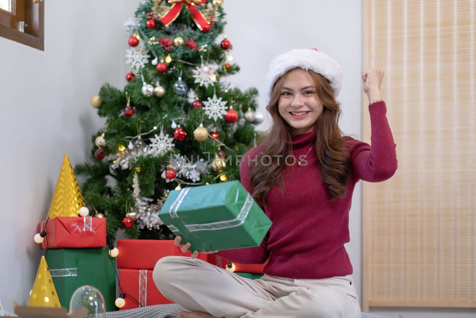 Cheerful asian young beautiful woman wearing santa hat showing open gift box with happy smiling face sitting in living room background...