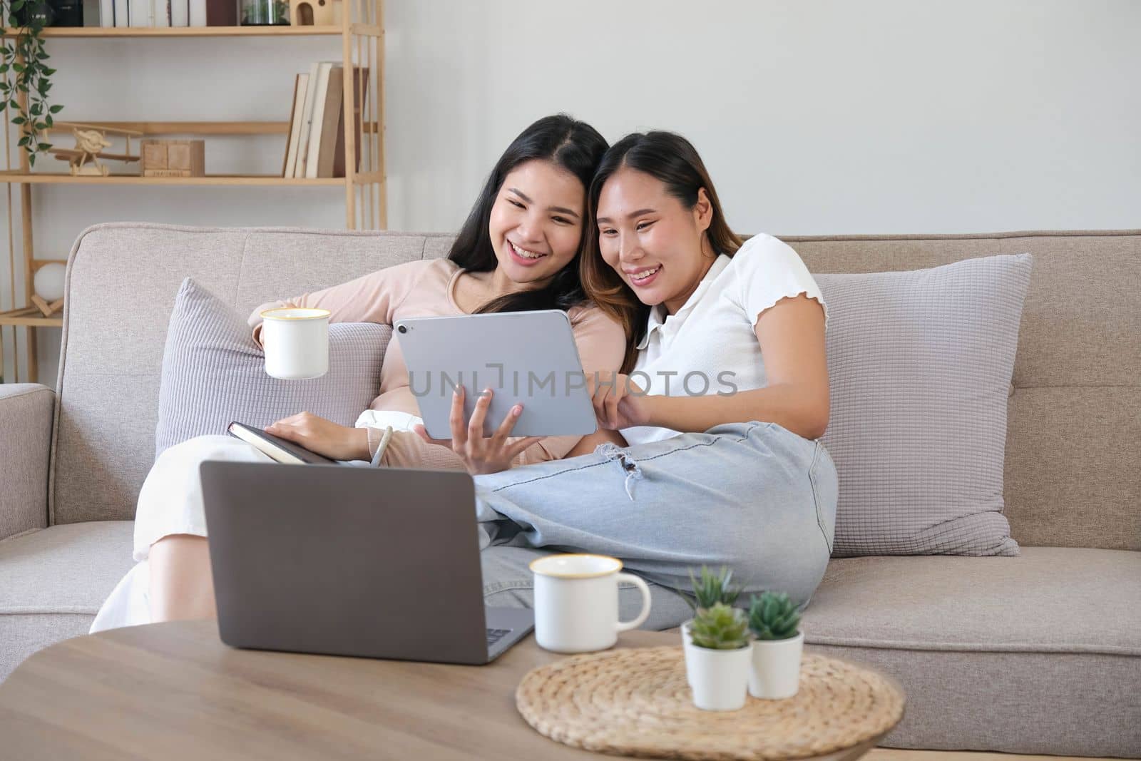 Two happy Asian women best friends in casual wear laughing while working with tablet at home in living room by wichayada