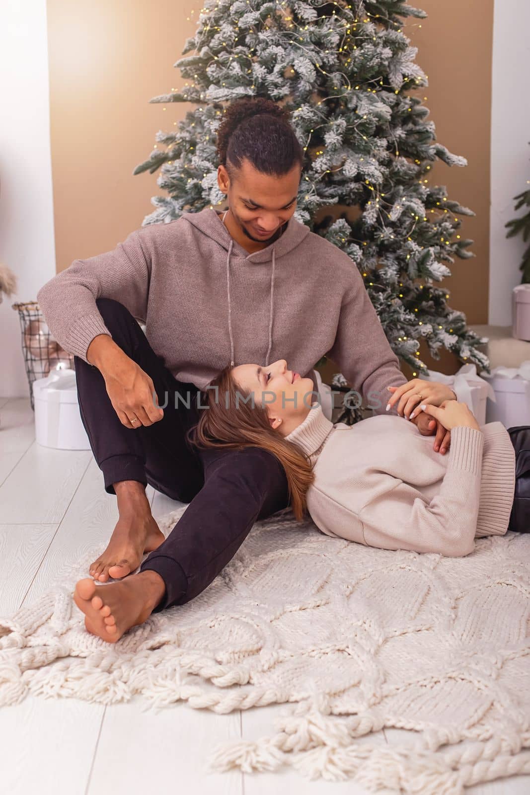 The girl, closing her eyes, lies on the lap of a stylish guy near the Christmas tree in the room.Vertical