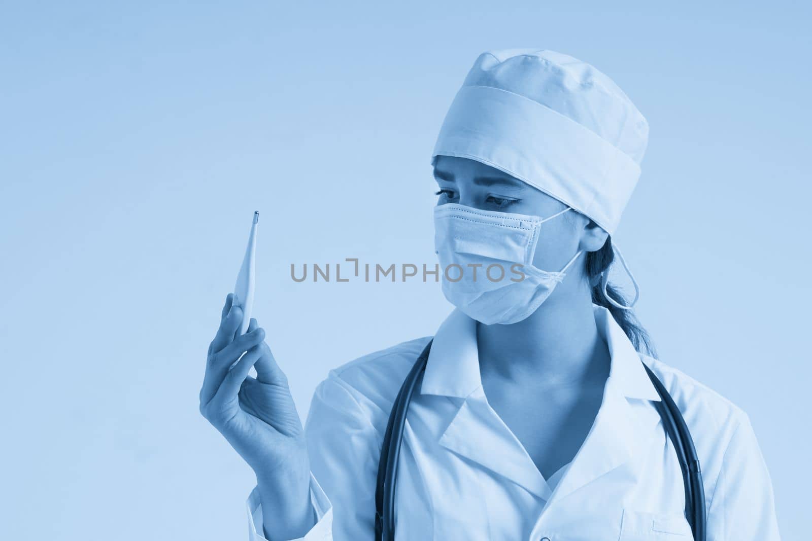 Doctor wearing face mask looking at measurement on medical thermometer over white background by Mariakray