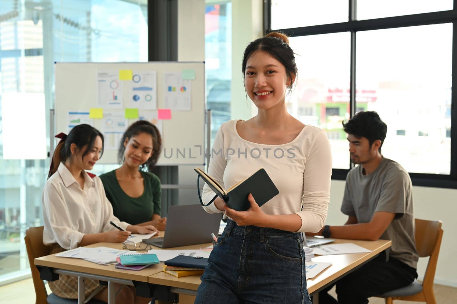 Positive young female team leader standing in front of meeting table at creative office and smiling to camera.