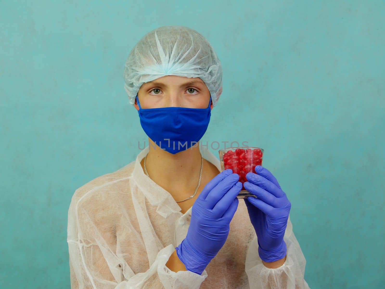 A young man wearing a medical mask shows off a glass of red pills. Studio shot indoors against a blue background. by gelog67