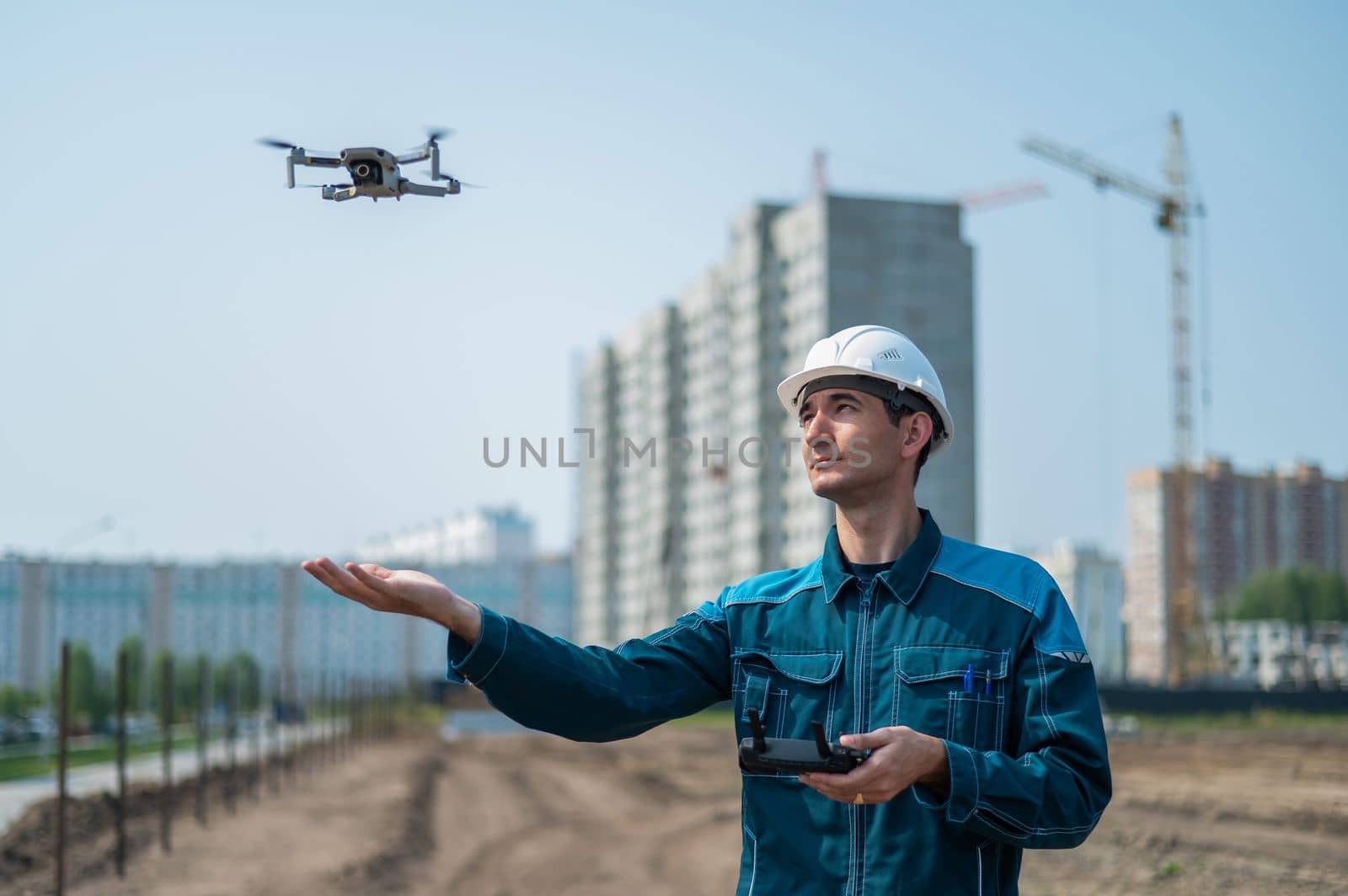 A man in a helmet and overalls controls a drone at a construction site. The builder carries out technical oversight