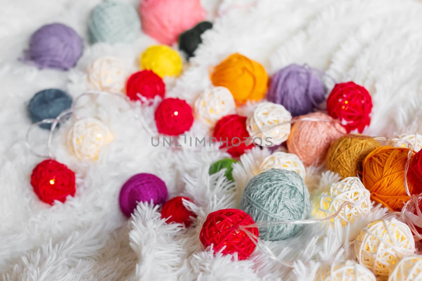 A lot of multicolored knitting yarn and a luminous garland in the form of balls lie on a white fluffy blanket. Cozy still life made of yarn. Close up. Copy space