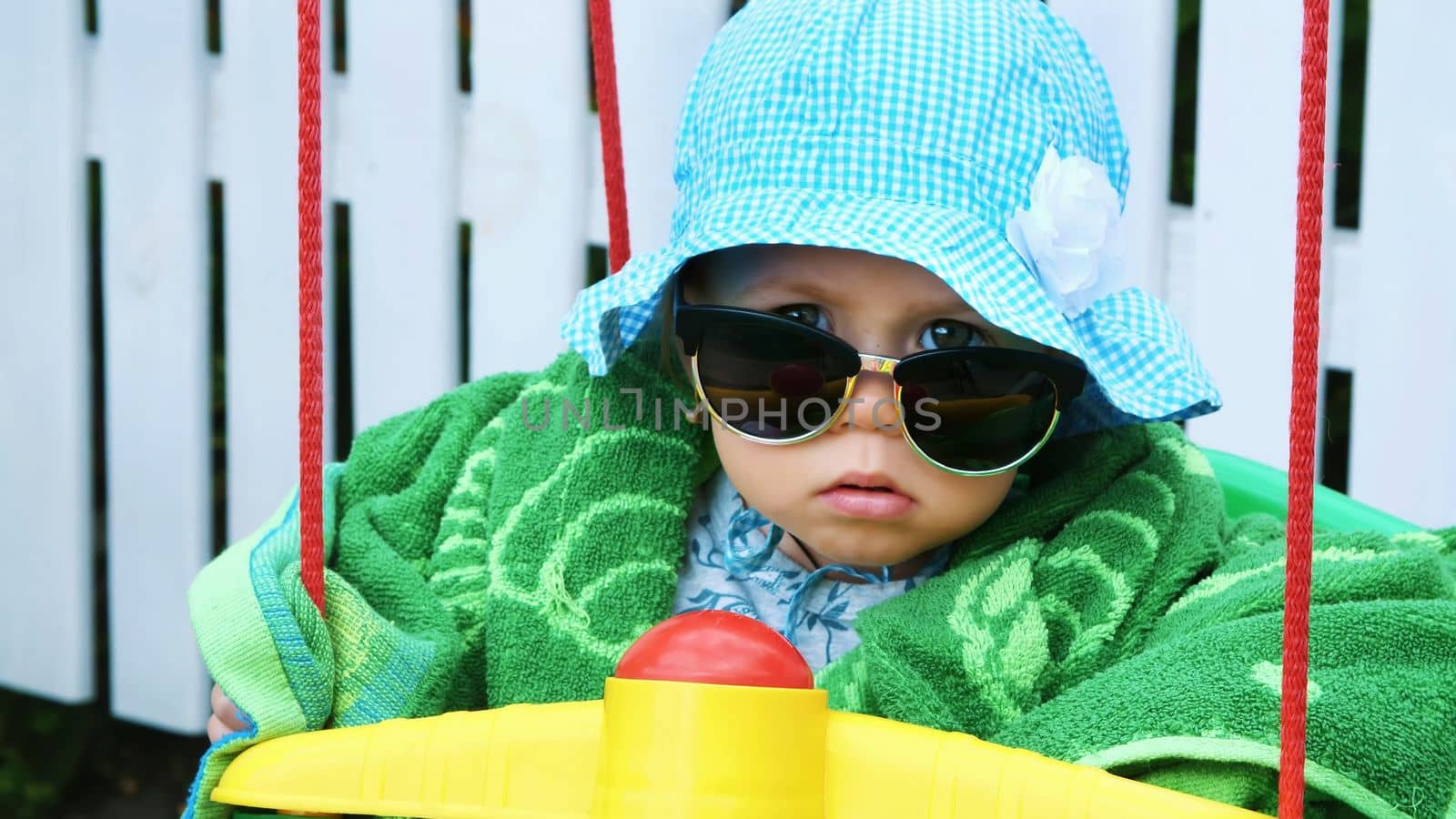 a one-year-old girl in a panama and sunglasses, like an important lady, sits in the children's swing and swings, in the summer, in the garden. High quality photo