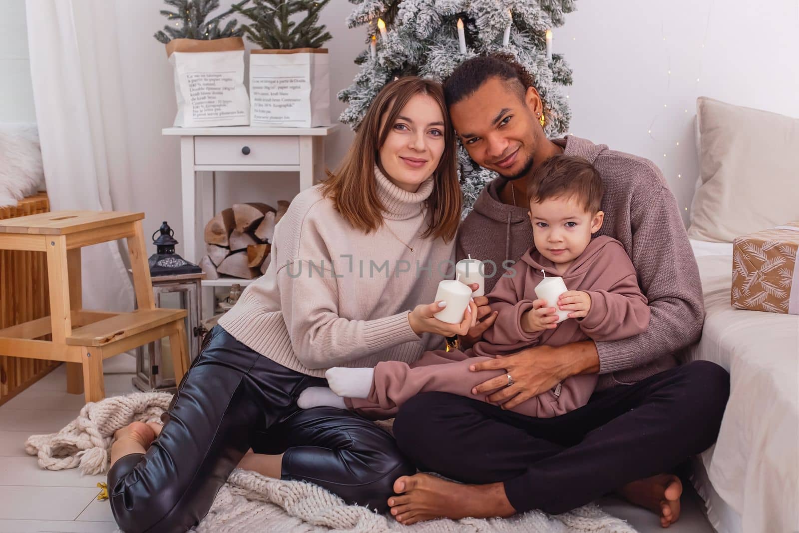 A young diverse family with a baby boy is sitting with candle near a Christmas tree by Zakharova