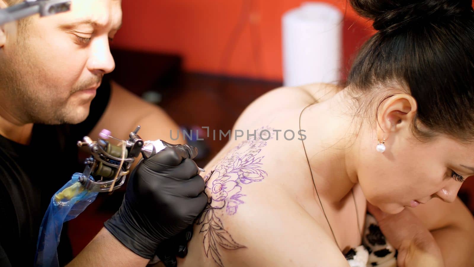 in a tattoo salon , a specialist is doing a tattoo on woman's back, a floral ornament. a man works in special gloves, on special equipment. drawing is done with black paint. High quality photo