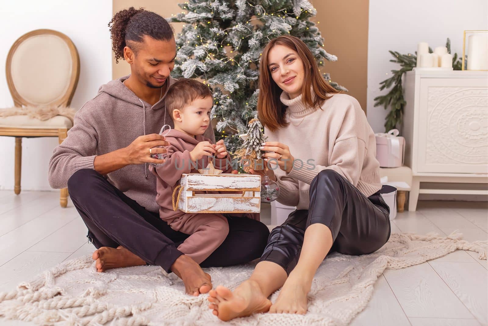 A happy multi-racial family sitting on a knitted blanket near the Christmas tree, smiling and holding a wooden box with Christmas toys.