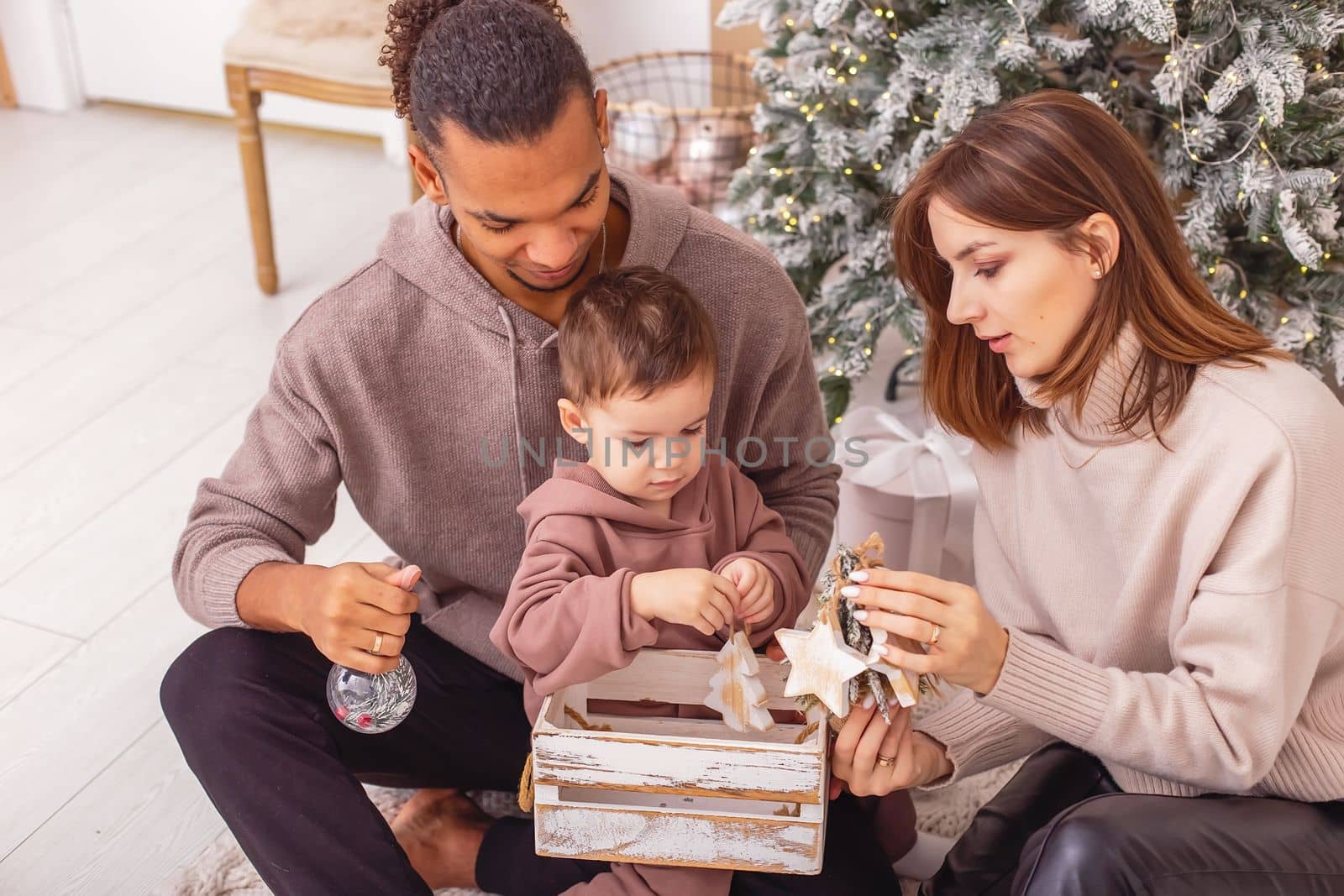 A beautiful stylish multi-racial family with a little boy near the Christmas tree, holding a wooden box with Christmas toys. Top view