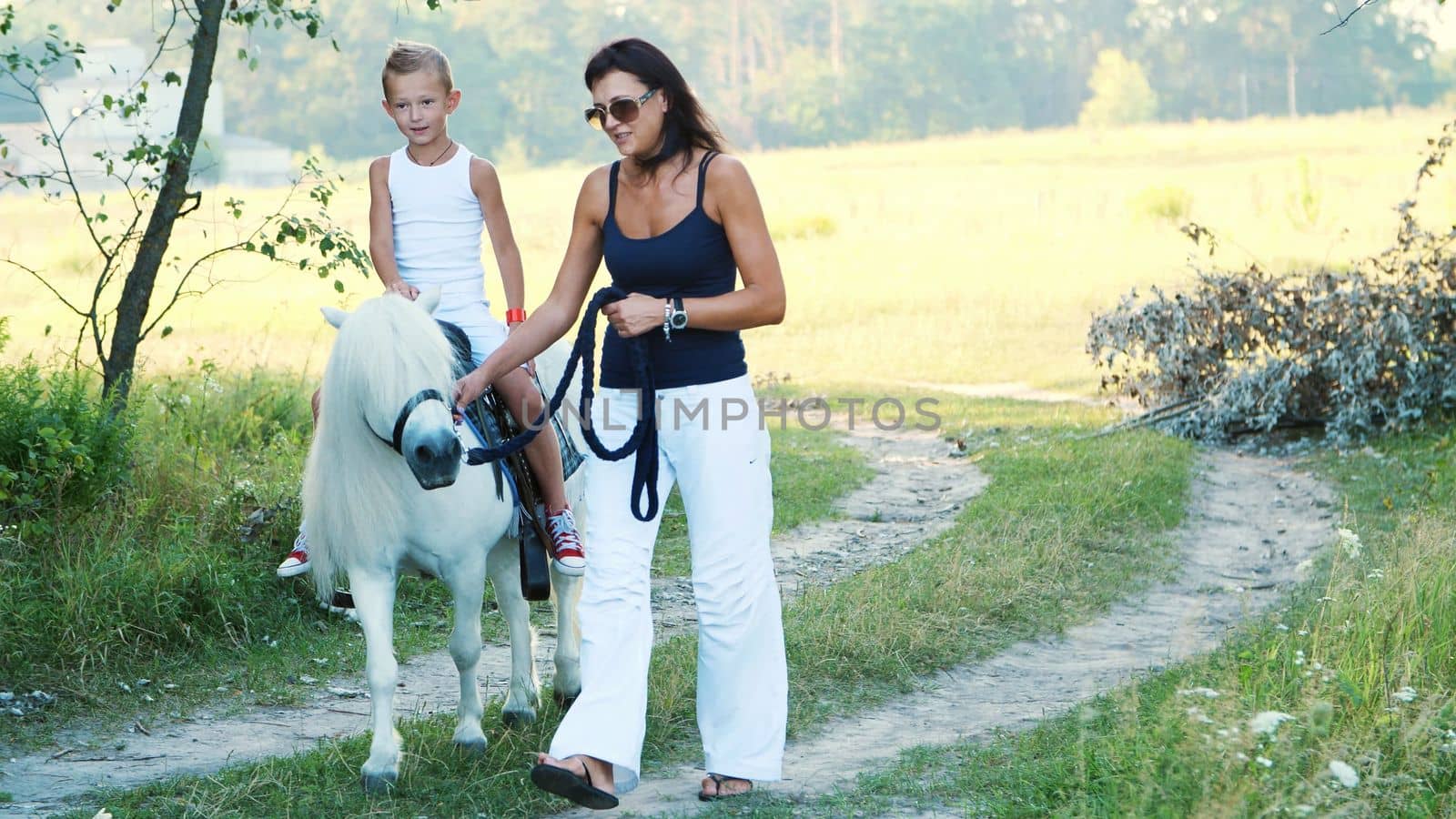 Mom and daughter are walking around the field, daughter is riding a pony, mother is holding a pony for a bridle. Cheerful, happy family vacation. Outdoors, in summer, near the forest. High quality photo