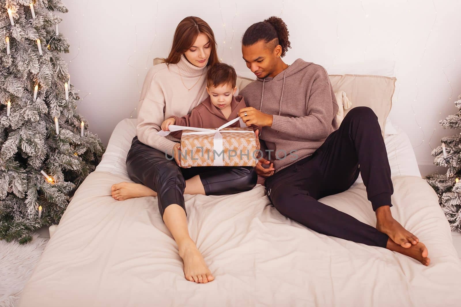 A stilysh multi-racial family is sitting on a bed, in a Christmas interior, untying a ribbon on a brown gift box