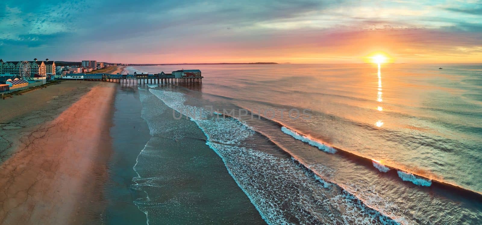 Image of Wood pier, beach, and shops with ocean waves from above during sunrise light